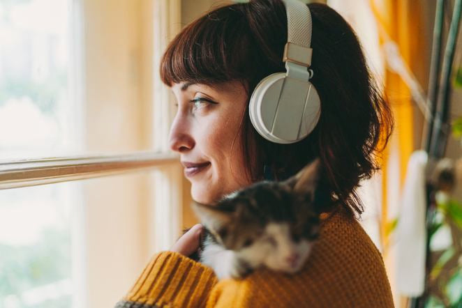 Frau mit Kopfhörern hält Katze am Fenster, warmes Tageslicht