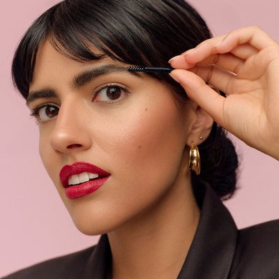 Frau bürstet ihre Augenbraue mit einem Bürstchen vor rosa Hintergrund