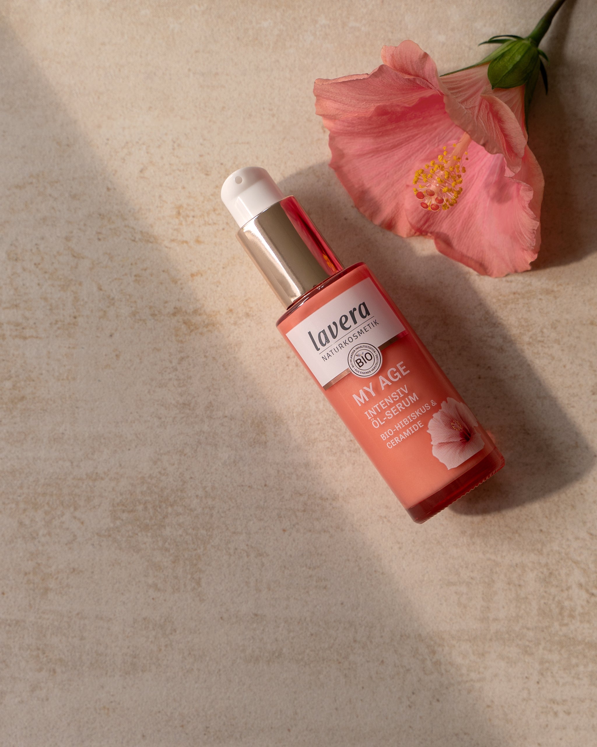 lavera MY AGE Intensiv Öl-Serum mit Bio-Hibiskus und Ceramiden, inszeniert neben einer rosa Hibiskusblüte auf heller Steinoberfläche.