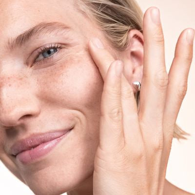 Eine blonde Frau streicht mit einem Finger über ihre Wange und lächelt.