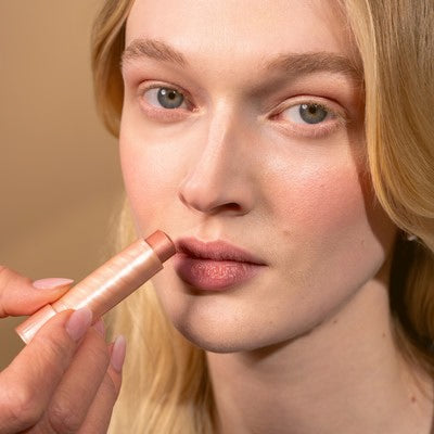 Junge Frau mit blonden Haaren, die in die Kamera lächelt und sich Multi Balm auf die Lippen aufträgt