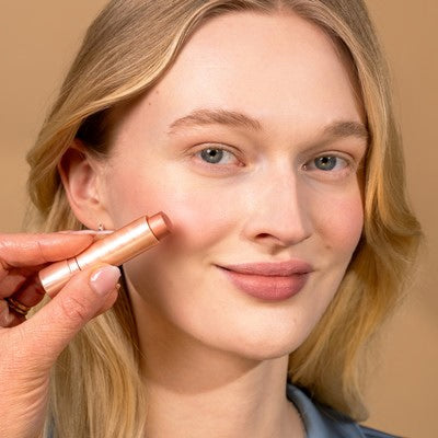 Junge Frau mit blonden Haaren, die in die Kamera lächelt und sich Farbe auf die Wange aufträgt